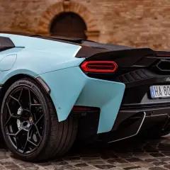 Niebieskie, nowe Lamborghini z czarnym spojlerem i felgami, zaparkowane na bruku przed ceglanym budynkiem.