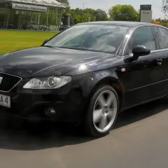 Czarny Seat Exeo 2.0 TDI w ruchu, z widocznym salonem Audi w tle.