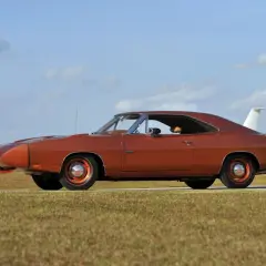 Pomarańczowy Dodge Daytona z charakterystycznym spojlerem i białym pasem z napisem "DAYTONA" na błotniku, zaparkowany na tle nieba z chmurami.