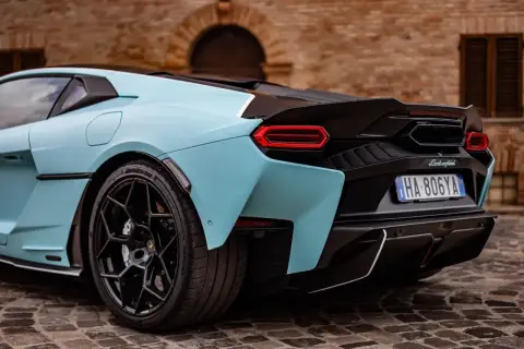 Niebieskie, nowe Lamborghini z czarnym spojlerem i felgami, zaparkowane na bruku przed ceglanym budynkiem.