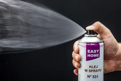 Dłoń trzyma puszkę z klejem w sprayu, który jest rozpylany. Idealny klej do podsufitki bez zdejmowania.