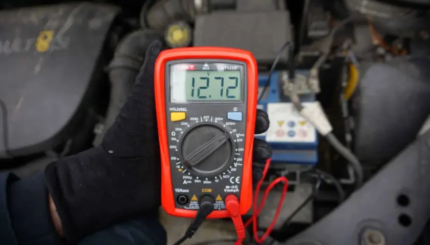 Ręka w czarnej rękawiczce trzyma multimetr pokazujący 12.72V, co pomaga sprawdzić akumulator.
