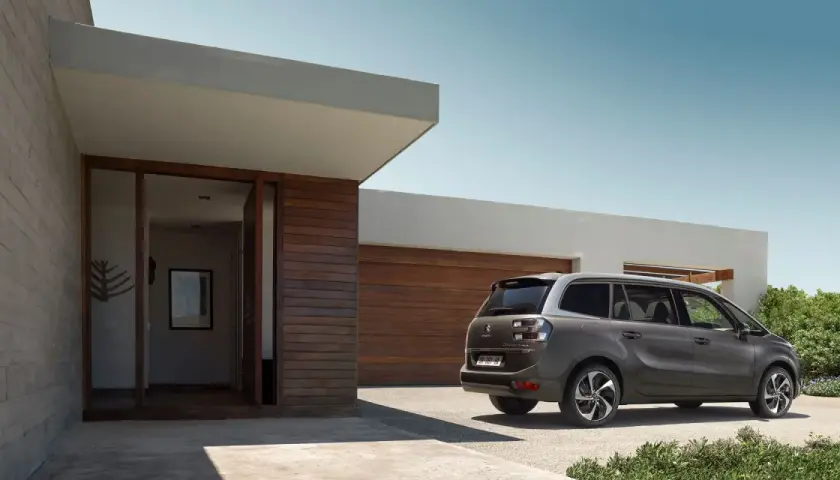 Szary minivan Citroën C4 Picasso zaparkowany przed nowoczesnym domem z drewnianą elewacją.