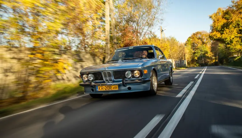 Błękitne BMW E9 z kierowcą jedzie jesienną drogą wśród kolorowych drzew.