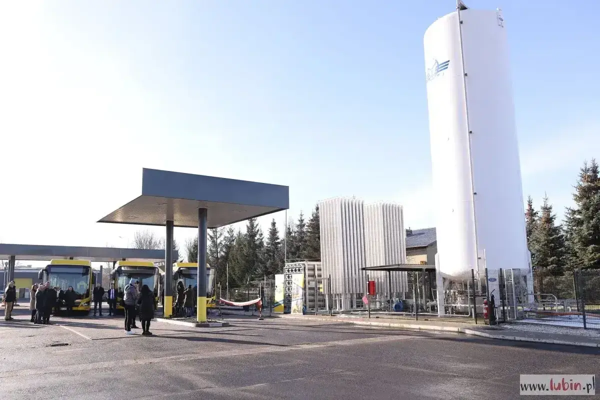 Stacja tankowania CNG. Żółte autobusy czekają pod wiatą, obok wysoki zbiornik i instalacja.