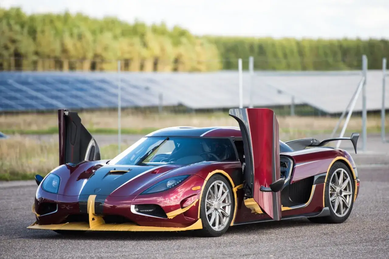 Czerwony Koenigsegg Agera RS z otwartymi drzwiami, gotowy do jazdy po torze z panelami słonecznymi w tle.