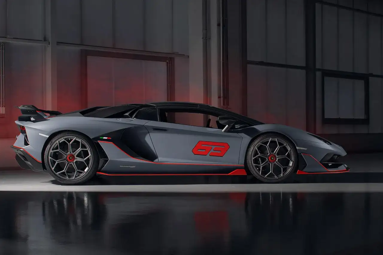 Szary Lamborghini Aventador z czerwonymi akcentami i numerem 63 na drzwiach, stojący w garażu z czerwonym światłem w tle.