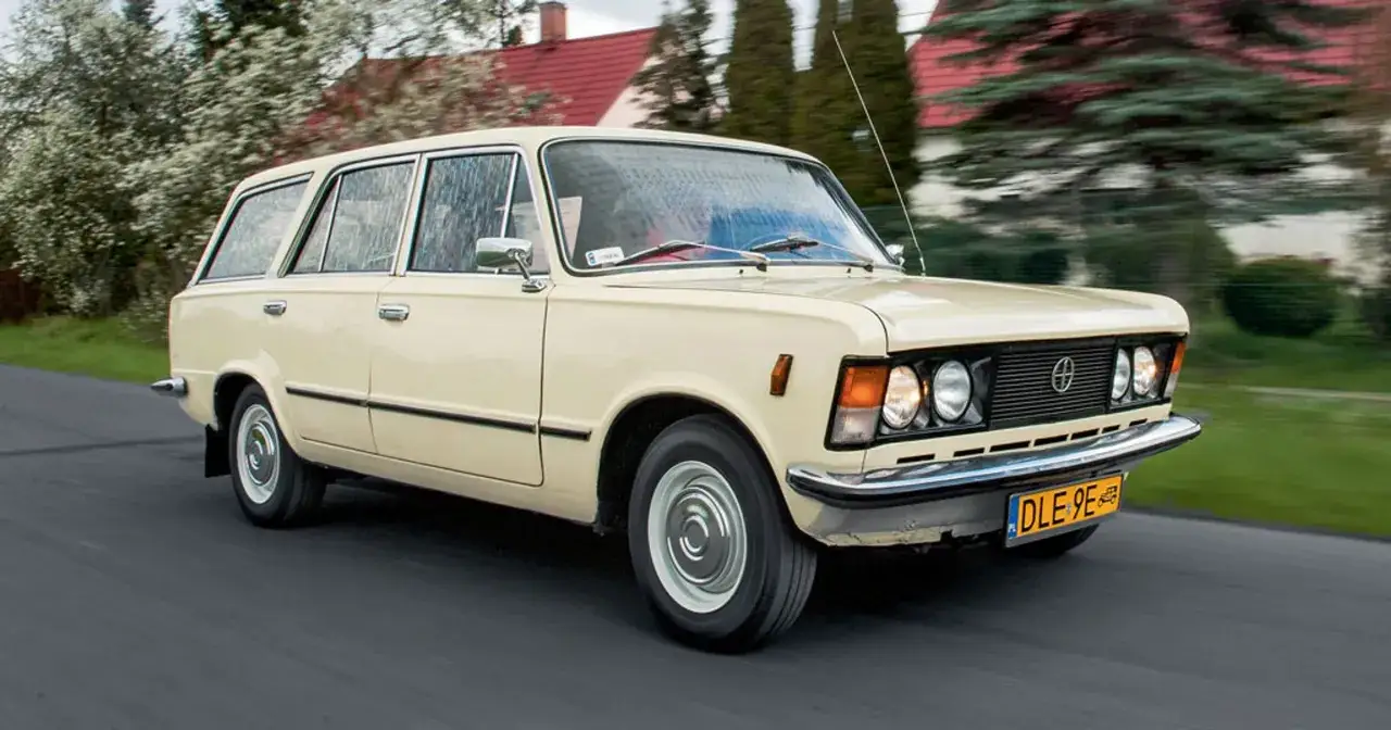 Kremowe kombi auto z lat 70. jedzie po drodze, w tle domy i drzewa.