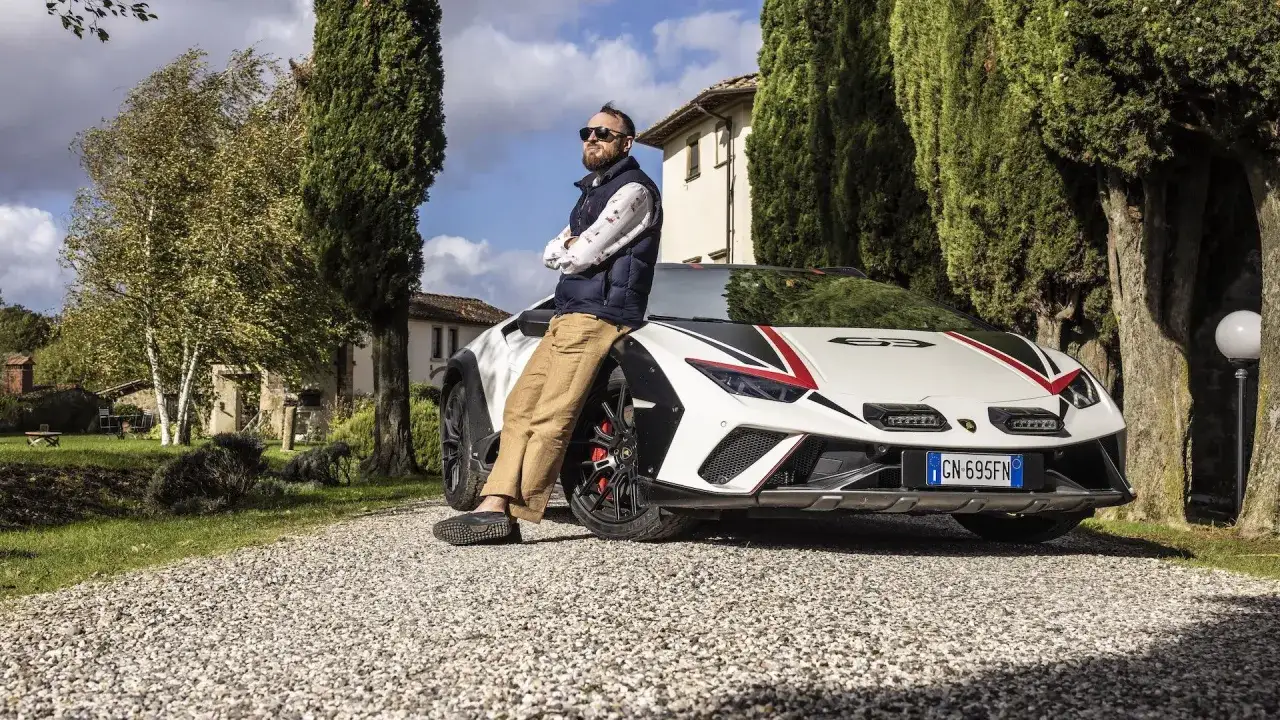 Mężczyzna w okularach przeciwsłonecznych opiera się o białe Lambo Huracan z czerwono-czarnymi akcentami, na tle włoskiej willi i cyprysów.