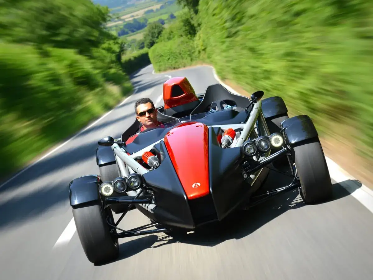 Mężczyzna w okularach przeciwsłonecznych prowadzi czerwono-czarny Ariel Atom po krętej drodze, otoczonej zielenią.